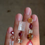 A close-up of a hand displaying three strands of gemstone beads. The beads include clear rectangular and round stones, a bright orange oval stone, deep red round stones, a yellow faceted stone, and a large pink teardrop stone.