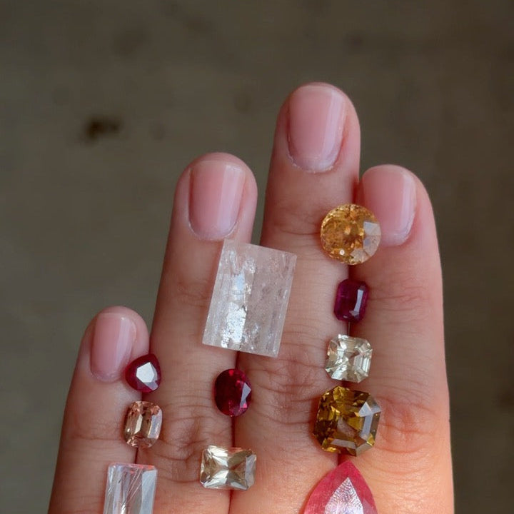 A close-up of a hand displaying three strands of gemstone beads. The beads include clear rectangular and round stones, a bright orange oval stone, deep red round stones, a yellow faceted stone, and a large pink teardrop stone.
