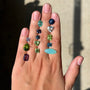 A hand displaying two vertical strands of colorful gemstone drop earrings against a sunlit beige wall.