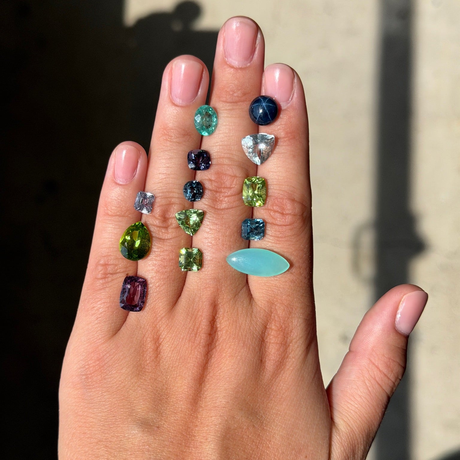 A hand displaying two vertical strands of colorful gemstone drop earrings against a sunlit beige wall.